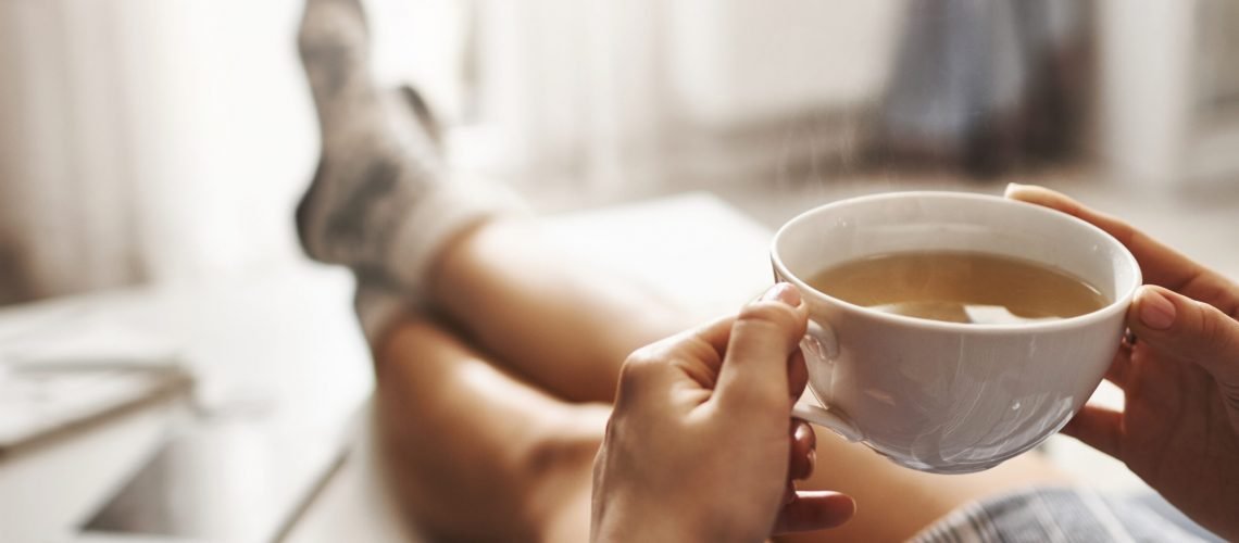 Cup of tea and chill. Woman lying on couch, holding legs on coffee table, drinking hot coffee and enjoying morning, being in dreamy and relaxed mood. Girl in oversized shirt takes break at home.